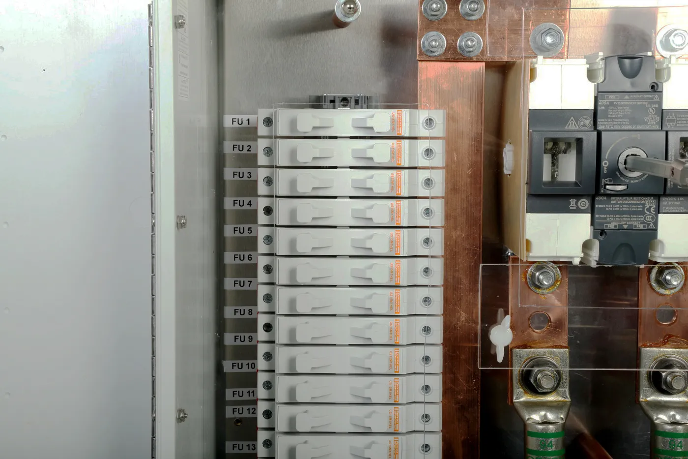 Close-up of labeled fuse holders FU 1 to FU 13 beside a DC disconnect switch and copper bus bars